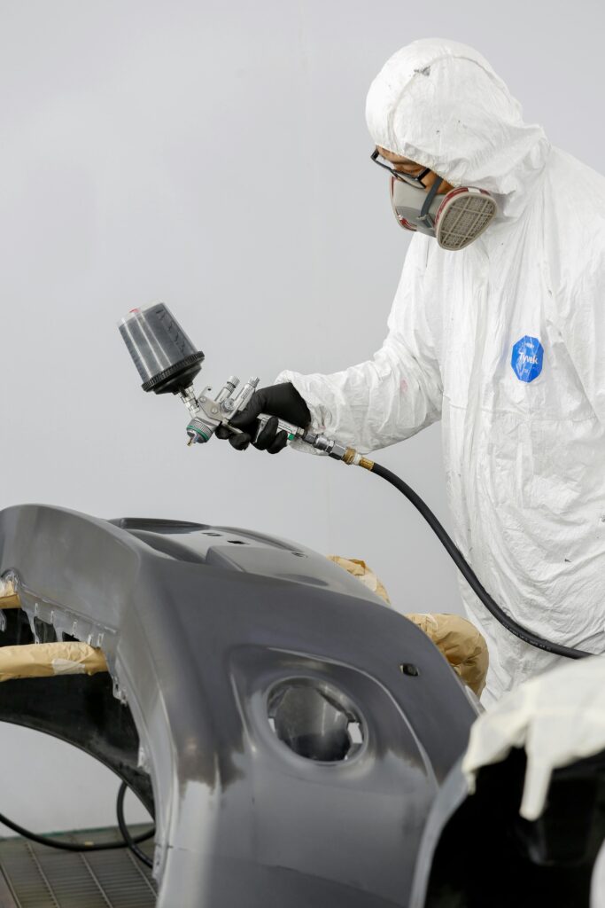 Technician in protective gear spray painting a car bumper.